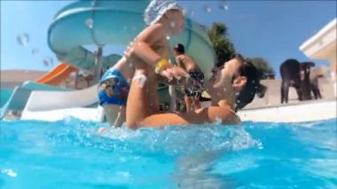 Ağır çekim görüntüleri oynamak ve onun yürümeye başlayan çocuk yüzme havuzu, aquapark atma mutlu gülümseyen Genç Anne