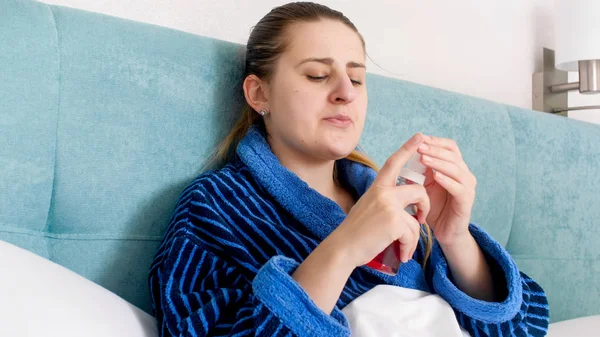 Closeup portrait of young woman feeling unwell lying in bed and holding spray from sore throat — Stock Photo, Image
