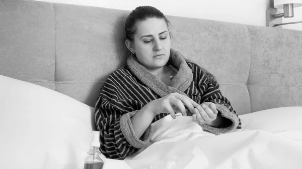 Retrato en blanco y negro de una joven enferma acostada en la cama y vertiendo pastillas en la mano de una botella de vidrio —  Fotos de Stock