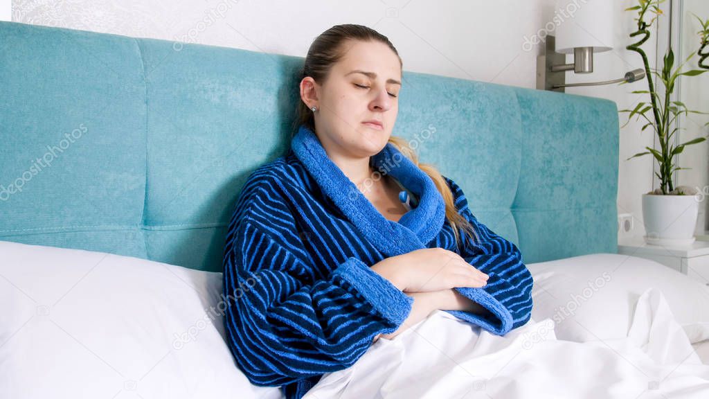 Portrait of young sick woman lying in bed and holding digital thermometer under arm