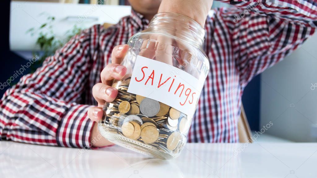 Closeup image of young man trying to get coins out of glass jar with family savings