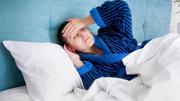 Retrato de una joven enferma en bata de baño acostada en la cama y con dolor de cabeza —  Fotos de Stock