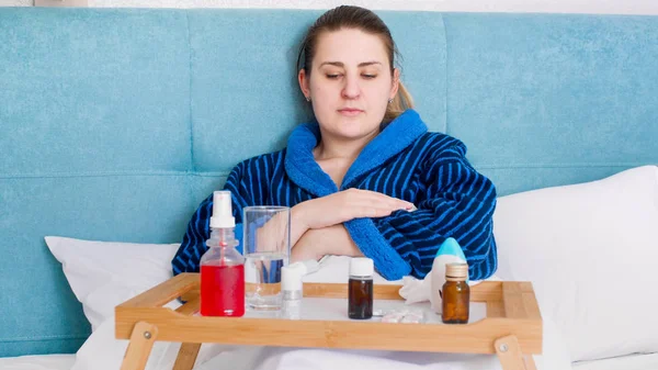 Portrait of sick young woman with fever lying in bed with tray full of medicines — Stock Photo, Image