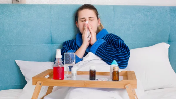 Portrait of sick young woman lying with lots of medicines and pills in bed and blowing nose in tissue — Stock Photo, Image