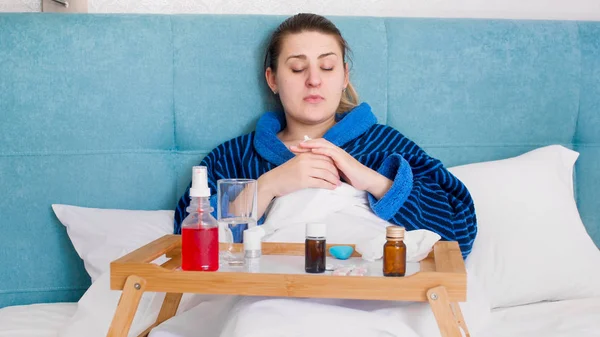 Портрет больной женщины в халате, лежащей в постели с большим количеством таблеток и лекарств на подносе — стоковое фото