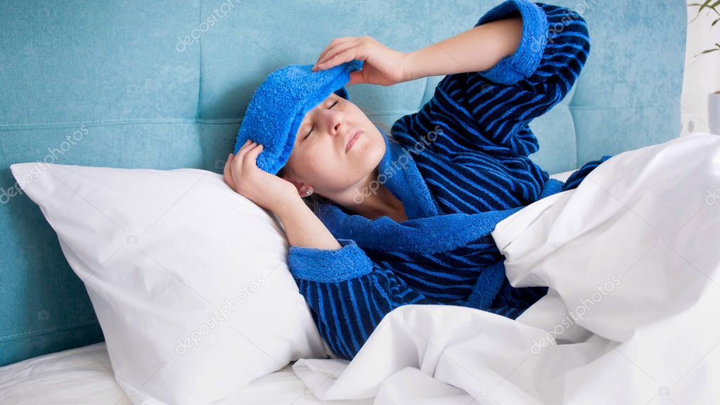 Portrait of young sick woman suffering from migraine putting wet cold towel on forehead