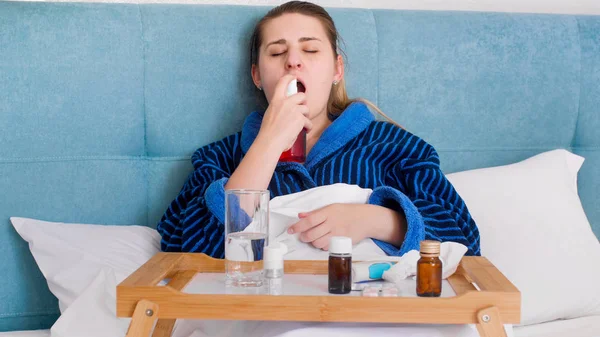 Portrait de jeune femme malade avec mal de gorge couchée au lit et utilisant un spray médicinal — Photo