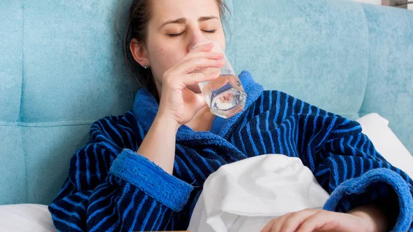 Retrato de una joven enferma acostada en la cama y bebiendo agua de un vaso — Foto de Stock