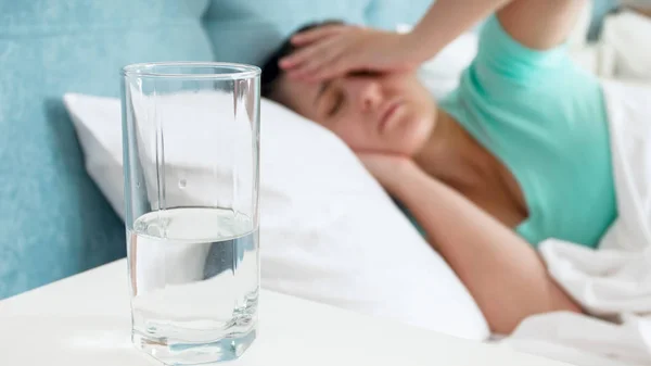 Immagine primo piano di vetro d'acqua contro donna con mal di testa sdraiata a letto — Foto Stock