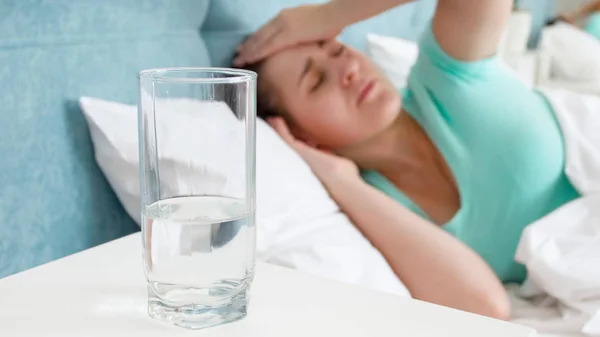 Imagen de la mujer enferma que sufre de dolor de cabeza acostada en la cama y vaso de agua en la mesita de noche —  Fotos de Stock