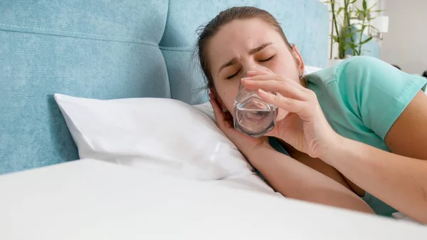 Portrait of young woman feeling unwell lying in bed and drinking water — Stock Photo, Image