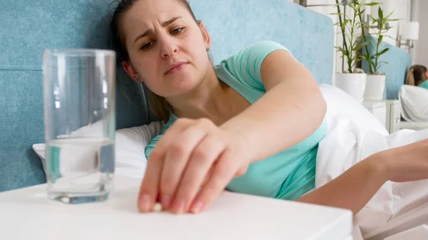 Портрет молодой больной женщины, лежащей в постели и принимающей таблетки с прикроватного столика — стоковое фото
