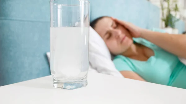 Closeup image of glass of water with aspirin on bedside table against woman with headache — Stock Photo, Image