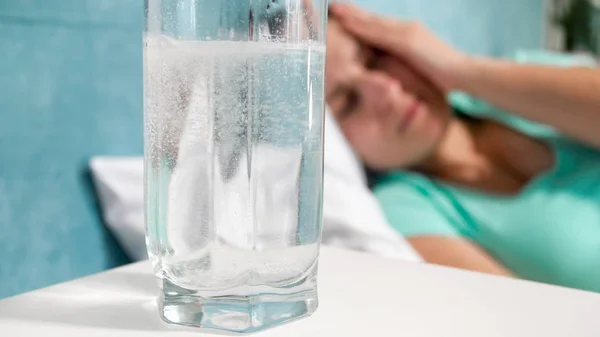 Image de verre d'eau avec aspirant et jeune femme souffrant de maux de tête couché dans le lit — Photo