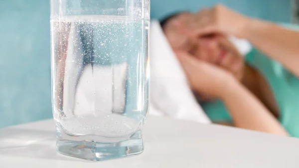 Imagen de primer plano del vaso de agua carbonatada en la mesita de noche contra la mujer que sufre de dolor de cabeza —  Fotos de Stock