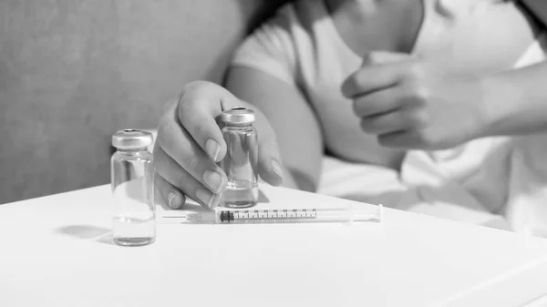 Image en noir et blanc d'une jeune femme allongée au lit prenant des ampoules avec des médicaments et une seringue sur la table de chevet — Photo
