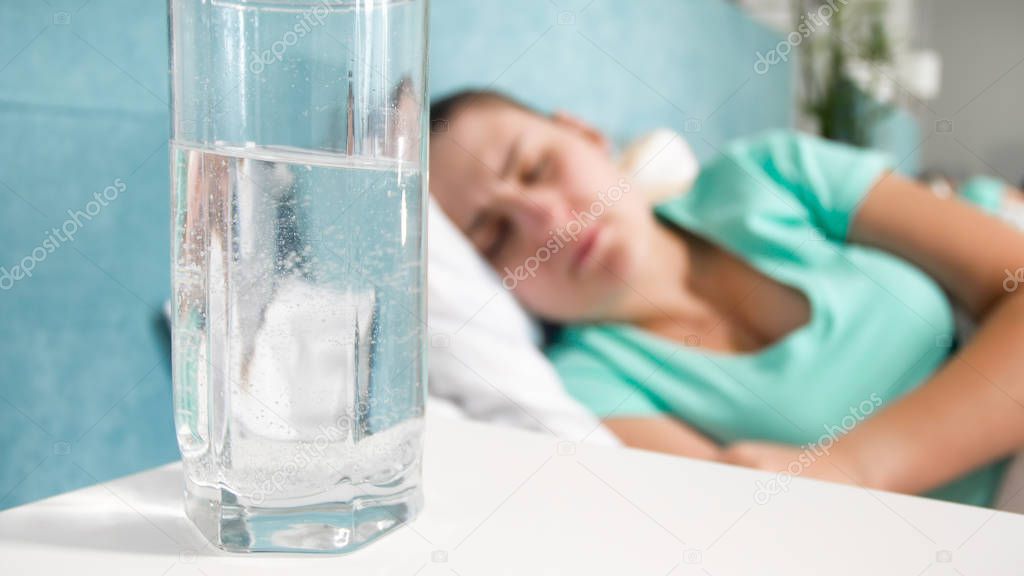 Closeup image of young woman feeling unwell lying in bed. Glass of water with aspirin on bedside table