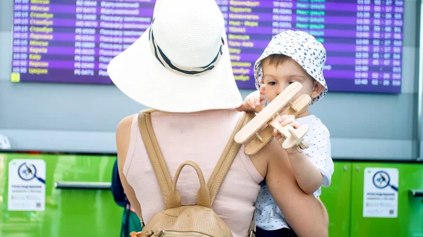 Ung kvinna håller hennes lille son och letar efter flygplanet avgångstid på skärmen i flygplats — Stockfoto