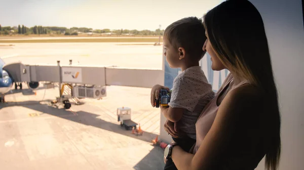 Imagem tonificada da jovem mãe segurando seu filho menor e mostrando aviões na pista através de uma grande janela no terminal do aeroporto — Fotografia de Stock