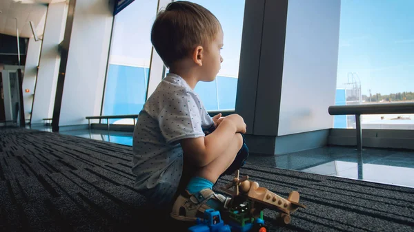 Симпатичный мальчик сидит на полу в аэропорту и смотрит в большое окно — стоковое фото