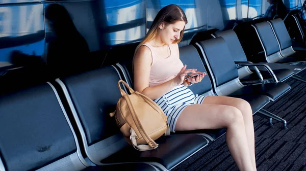 Schöne junge Frau sitzt im Wartezimmer am Flughafen-Terminal und benutzt Smartphone — Stockfoto