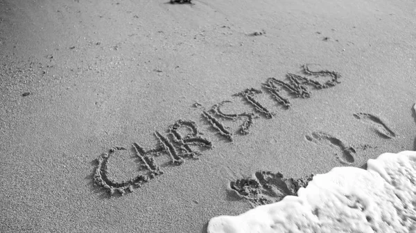 Black and white photo of word Christmas written on wet san on the beach. Sea wave flowing over. Concept of winter holidays, New Year and tourism — Stock Photo, Image
