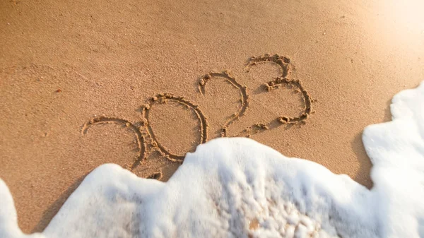 Närbild foto av havsvågor rullande över 2023 numrerar skriftligt på våt sand. Begreppet nyår, jul och resa på vintersemester. — Stockfoto