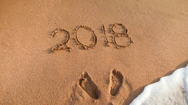 Närbild foto av havets vågor rulla över fotspår och 2018 tal skrivna på våta strand sand. Begreppet nyår, jul och resa på vintersemester. — Stockfoto