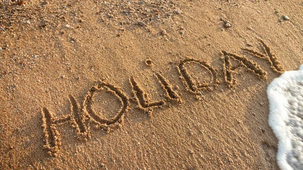 Zbliżenie zdjęcie fal morskich toczenia nad słowem wakacje napisane na piasku plaży. — Zdjęcie stockowe