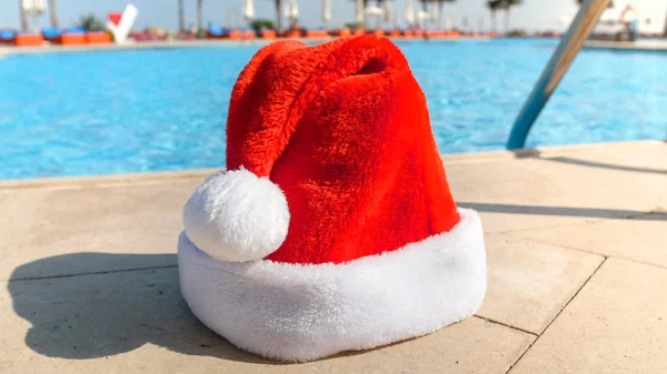 Closeup photo of Santa left his hat at the swimming pool on sunny day. Concept of travel and tourism on Christmas, New Year and winter holidays. — Stock Photo, Image