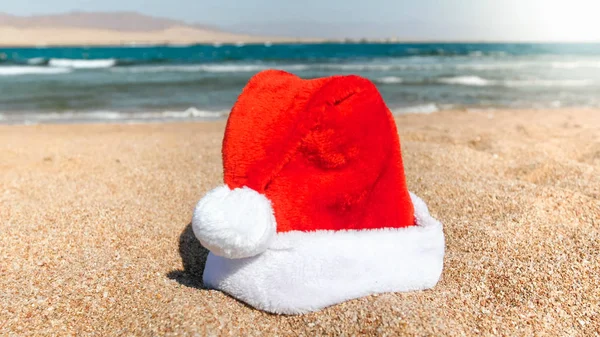 Imagem de close-up do chapéu Santas vermelho deitado na areia da praia do oceano. Conceito de viagem e turismo no Natal, Ano Novo e férias de inverno . — Fotografia de Stock