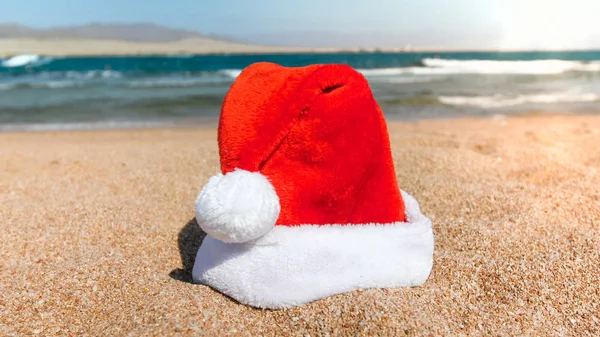 Foto close-up do chapéu de Papai Noel contra as ondas ea na praia no dia ensolarado. Conceito de viagem e turismo no Natal, Ano Novo e férias de inverno . — Fotografia de Stock