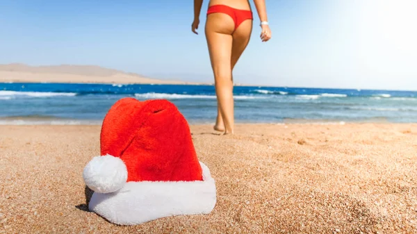 Close-up foto van Santa clauss hoed liggend op het strand tegen sexy vrouw in rode bikini. Concept van reizen en toerisme op kerst, Nieuwjaar en winter vakantie. — Stockfoto