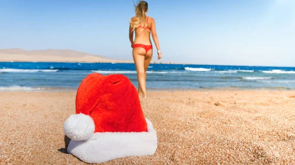 Giovane donna sexy in bikini rosso che cammina sulla spiaggia contro il cappello di Babbo Natale. Concetto di viaggio e turismo a Natale, Capodanno e vacanze invernali . — Foto Stock