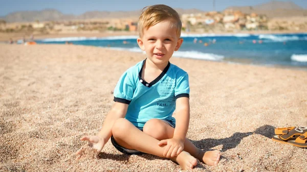 Szczęśliwy uśmiechnięty mały chłopiec w niebieskim t-shirt siedzi na plaży w jasnym słoneczny dzień i patrząc w aparacie — Zdjęcie stockowe