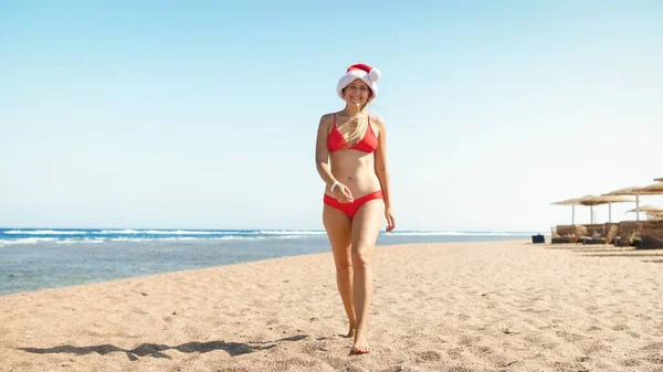 Beautiful laughing young woman in red Santa Claus hat walking on the sandy beach