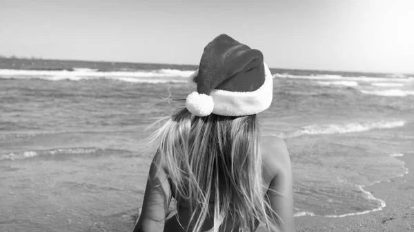 Imagem em preto e branco da jovem mulher usando chapéu Santas sentado na praia e olhando para o mar. Conceito de viagem e turismo no Natal, Ano Novo e férias de inverno . — Fotografia de Stock