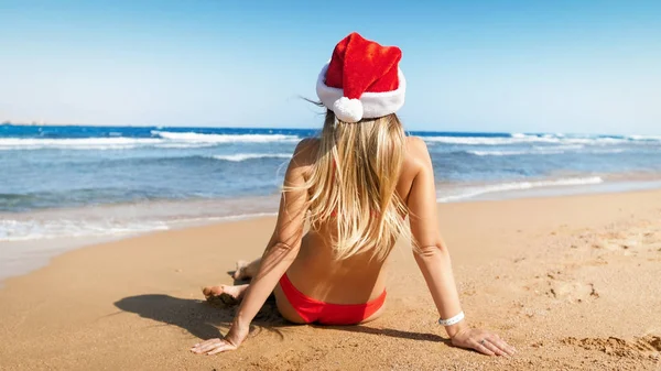 Achteruitkijk beeld van sexy jonge vrouw in kerstmuts en bikini zittend op het strand. Concept van reizen en toerisme op kerst, Nieuwjaar en winter vakantie. — Stockfoto