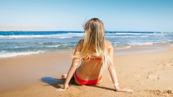 Rera view image of young woman in red bikini sitting on the beach and looking at sea — Stock Photo, Image