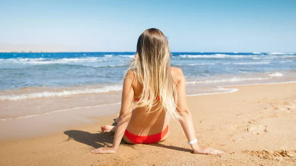 Sahilde oturan ve ufka bakarak uzun sarı saçlı seksi genç kadın — Stok fotoğraf