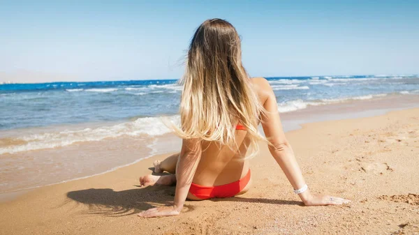 Sexy mujer joven con el pelo largo sentado en la arena y mirando al mar —  Fotos de Stock