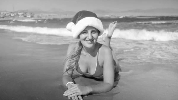 Ritratto in bianco e nero di bella donna sorridente in berretto Babbo Natale sdraiato sulla spiaggia del mare. Concetto di viaggio e turismo a Natale, Capodanno e vacanze invernali . — Foto Stock