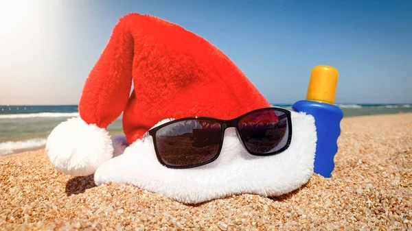Close-up beeld van de Kerstman muts met zonnebril en zonnebrand lotion liggend op het strand. Concept van reizen en toerisme op Kerstmis, Nieuwjaar en wintervakantie. — Stockfoto