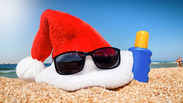 Photo rapprochée de la casquette du Père Noël avec des lunettes de soleil et de la crème solaire sur la plage lors d'une chaude journée ensoleillée. Concept de voyage et de tourisme à Noël, Nouvel An et vacances d'hiver . — Photo