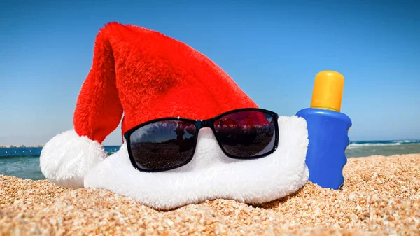 Foto di primo piano di occhiali da sole, cappello rosso di Babbo Natale e crema solare sdraiato sulla spiaggia di sabbia. Concetto di viaggio e turismo a Natale, Capodanno e vacanze invernali . — Foto Stock