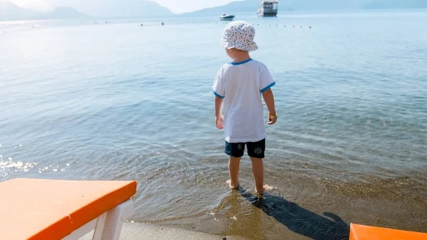 Widok z tyłu mały chłopiec maluch stojący w wodzie morskiej i patrząc z dala — Zdjęcie stockowe