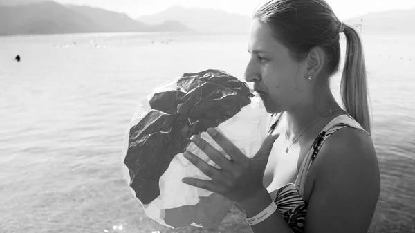 Black and white portrait of beautiful young woman blowing up colorful beach ball at sea — Stock Photo, Image