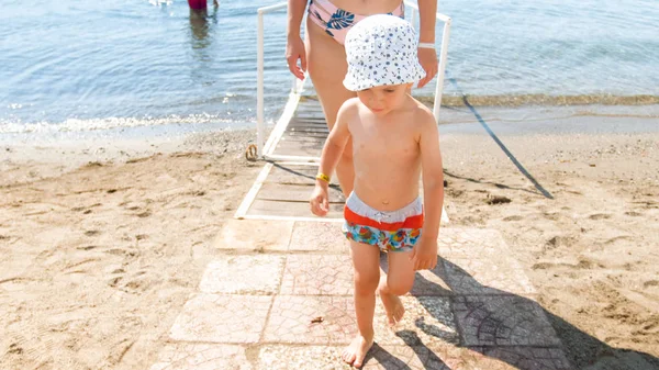 Uśmiechnięty mały chłopak w Kapelusz przeciwsłoneczny chodzenie z morza na piaszczystej plaży — Zdjęcie stockowe