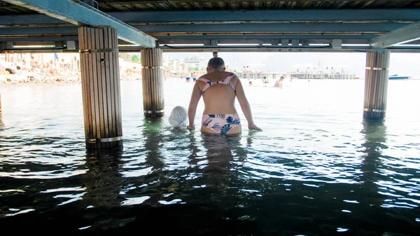 Giovane donna in costume da bagno che tiene il figlio bambino a mano e cammina sotto il molo di legno in mare — Foto Stock