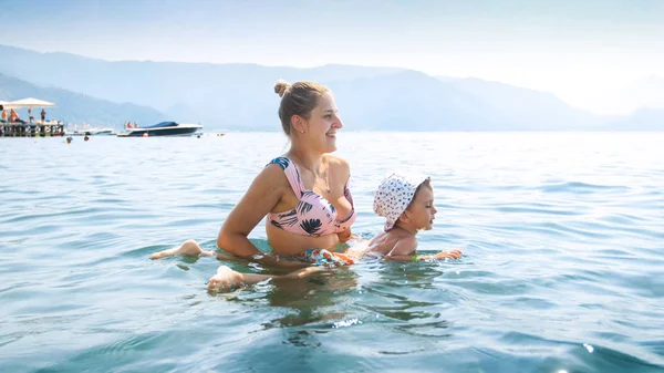Beautiful young woman holding her toddler child and helping him to swim in the sea — Stock Photo, Image